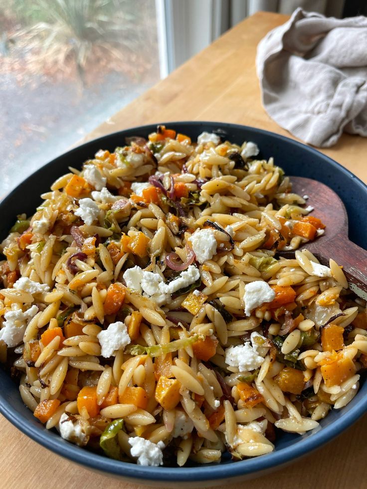 a blue bowl filled with pasta and vegetables on top of a wooden table next to a window