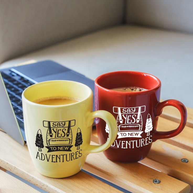 two coffee mugs sitting next to each other on top of a wooden table near a laptop