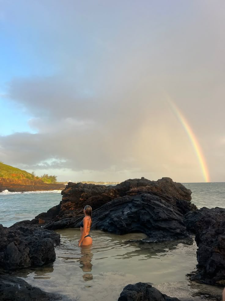 a person standing in the water with a rainbow in the sky behind them and rocks to the side