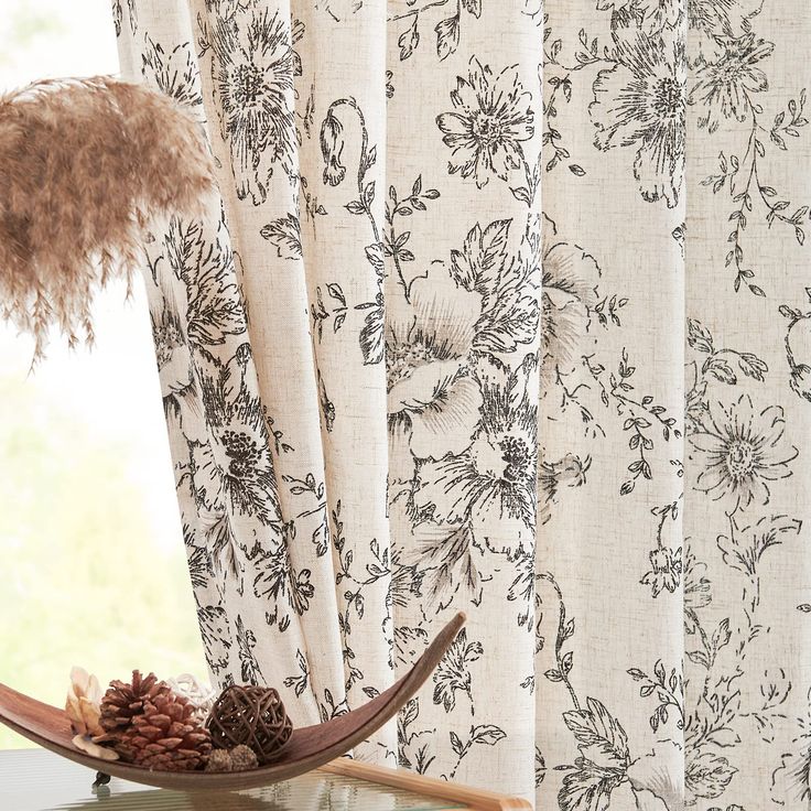 a window with floral curtains and pine cones on the table next to it, in front of an open window