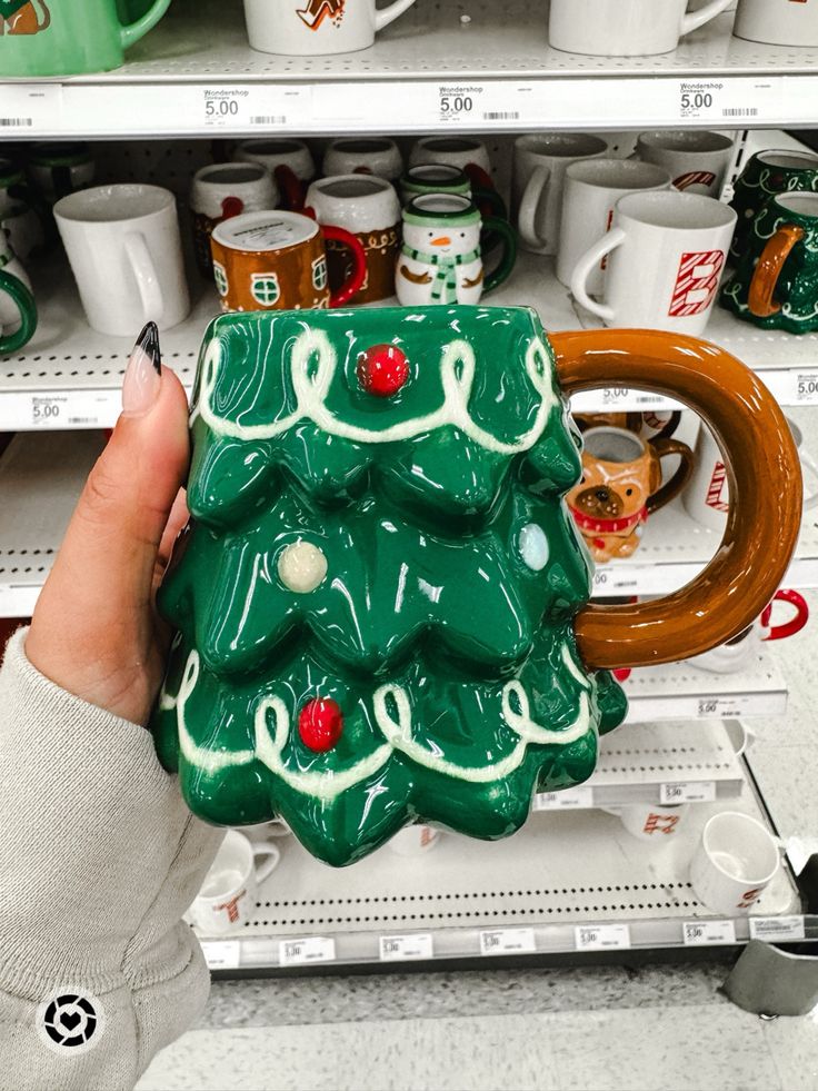 a hand holding a ceramic christmas tree mug in front of shelves with coffee mugs