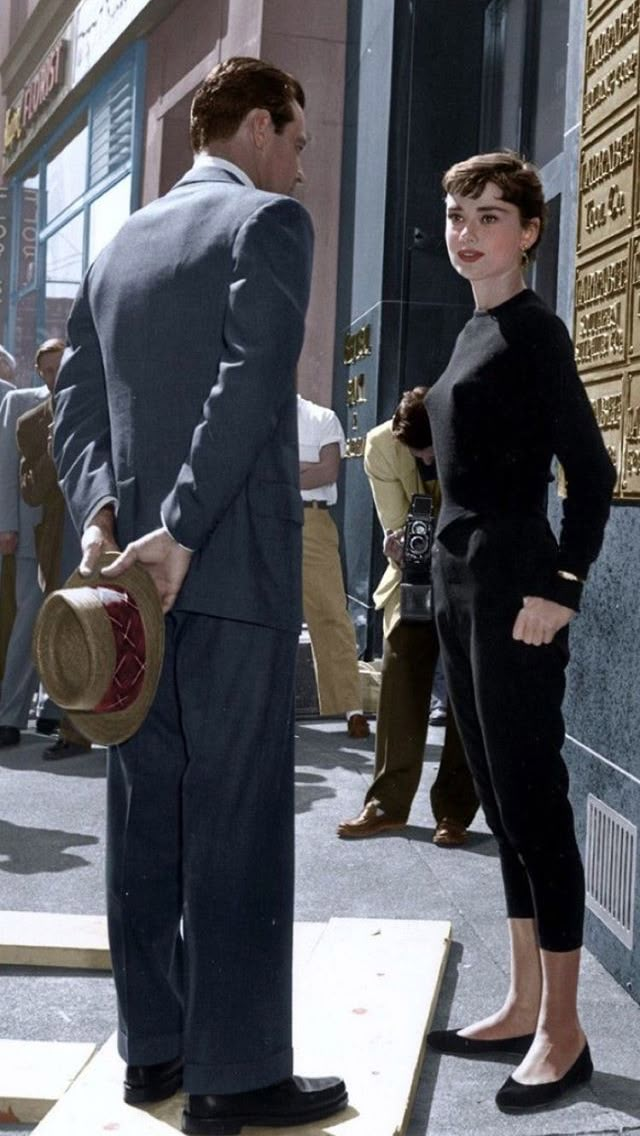 a man standing next to a woman in front of a building with a hat on her head