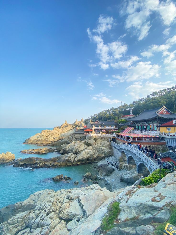 people are walking on the rocks near the water and some buildings in the distance with blue skies above them