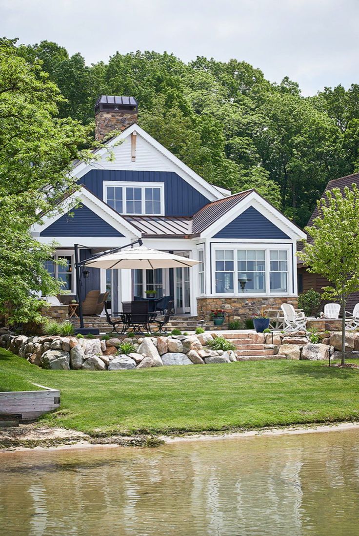 a blue house sitting on top of a lush green field next to a lake in front of a forest