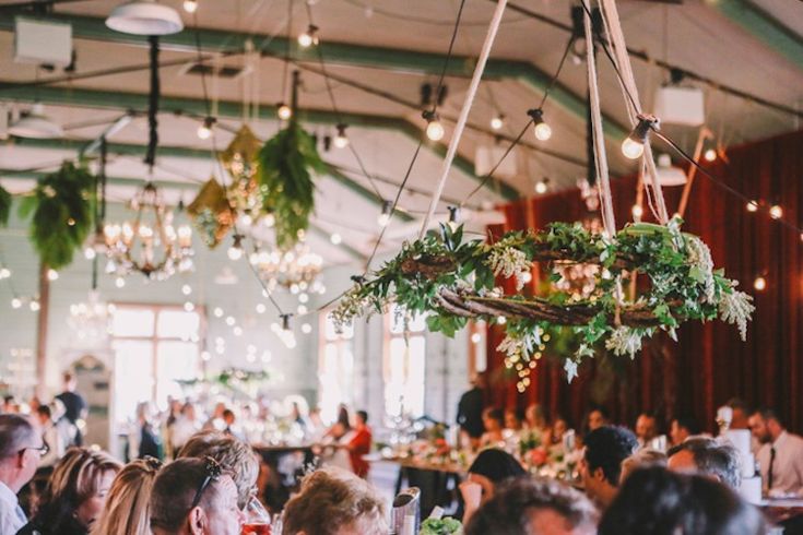 a large group of people sitting at tables in a room with plants hanging from the ceiling