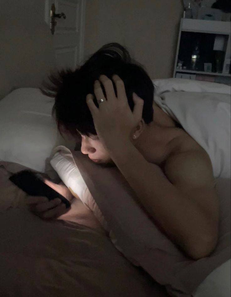 a shirtless man laying in bed with his hand on his head while looking at a cell phone