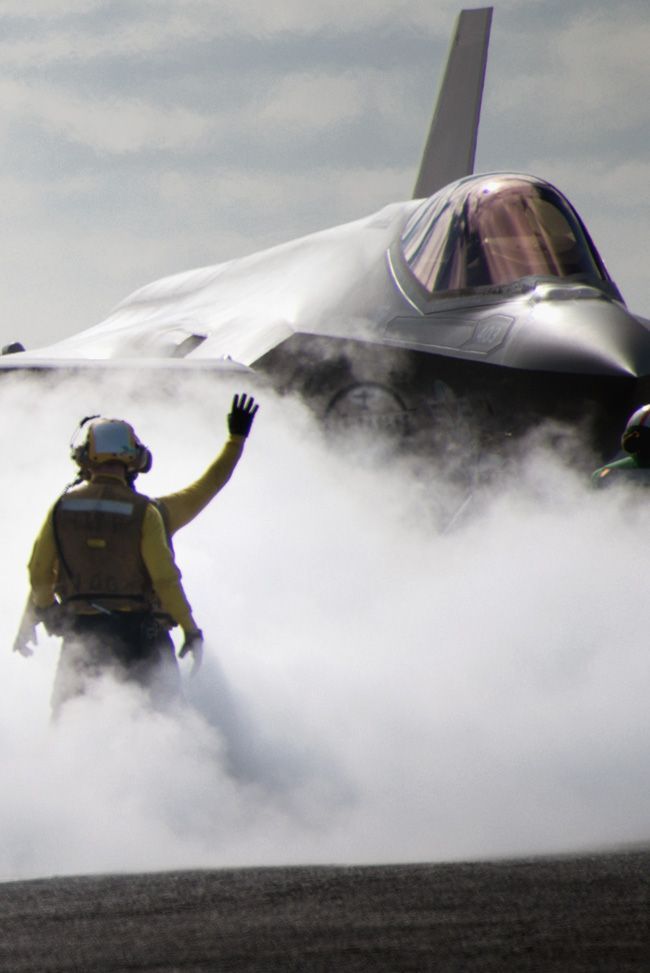 a fighter jet sitting on top of an airport runway next to a man in yellow jacket