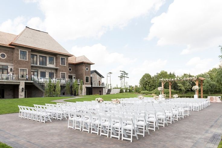 an outdoor wedding setup with white chairs and flowers on the aisle, in front of a large brick building