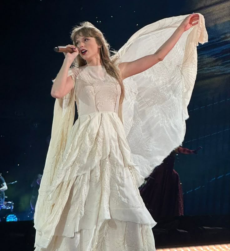 a woman in a white dress with wings on her shoulders and holding a microphone up to her mouth