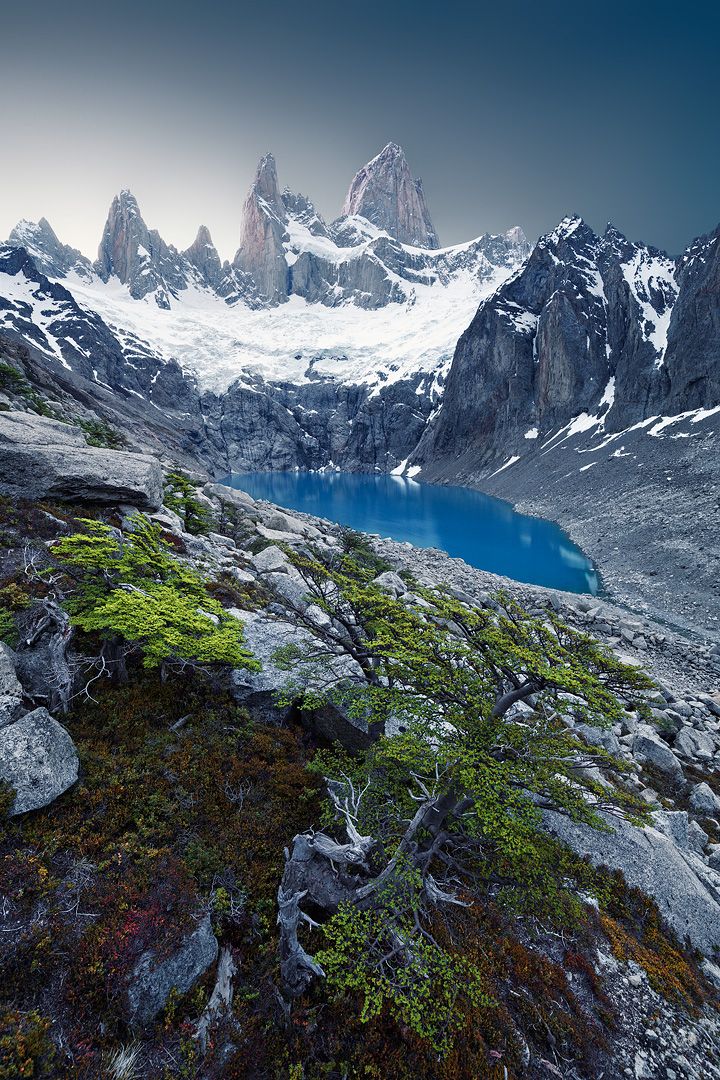 the mountains are covered in snow and green moss growing on it's sides, with a blue lake surrounded by rocks