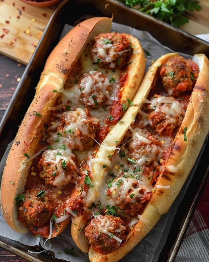 two hot dogs with meatballs and cheese on a baking tray next to a bowl of parsley