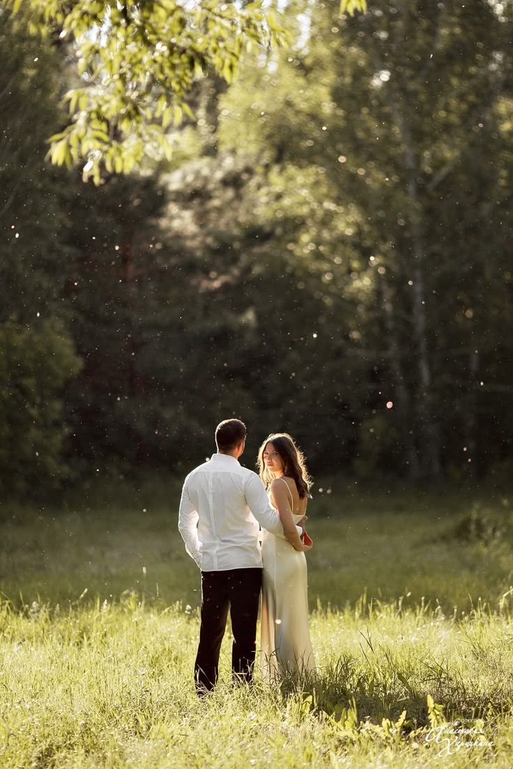 a man and woman standing in the middle of a grassy field with trees behind them