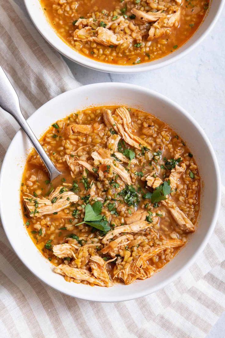 two bowls of chicken and barley soup with a spoon in it on a white table cloth