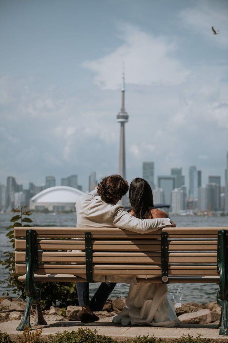 two people sitting on a bench in front of the water and a cityscape