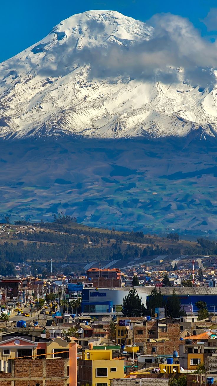 a large snow covered mountain towering over a small town in the foreground with buildings below