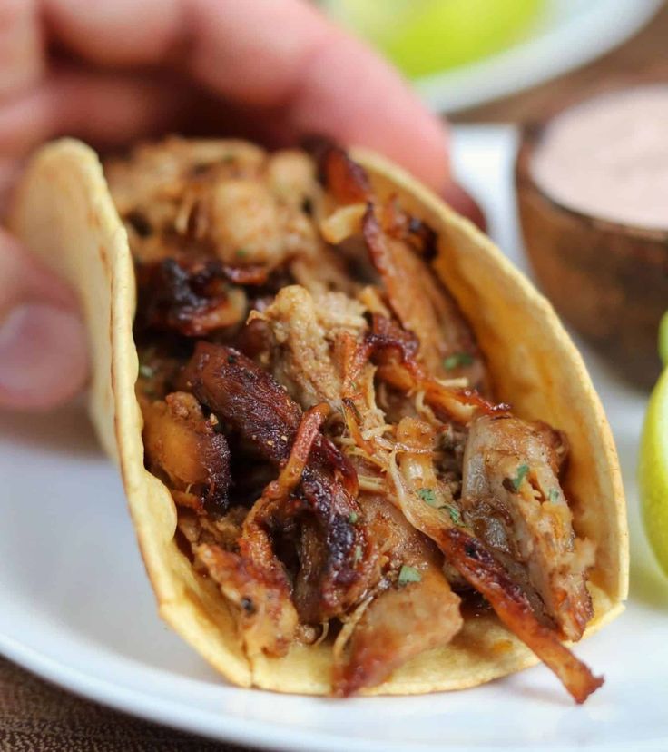 a person holding a taco on a white plate with limes and sauce in the background