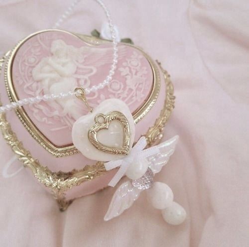 a pink heart shaped box sitting on top of a white cloth covered bed next to a beaded angel