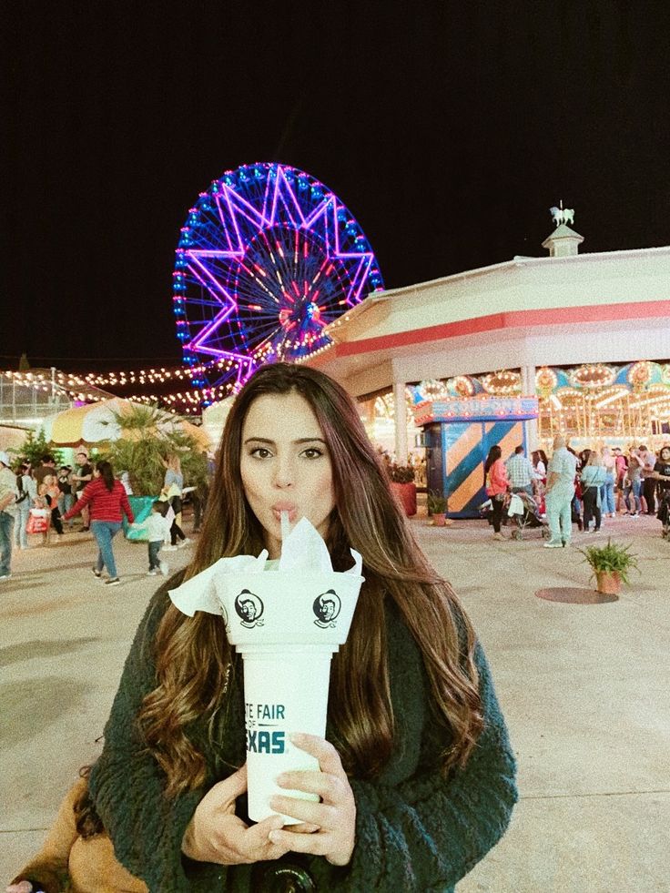 a woman standing in front of a ferris wheel at night holding up a paper cup