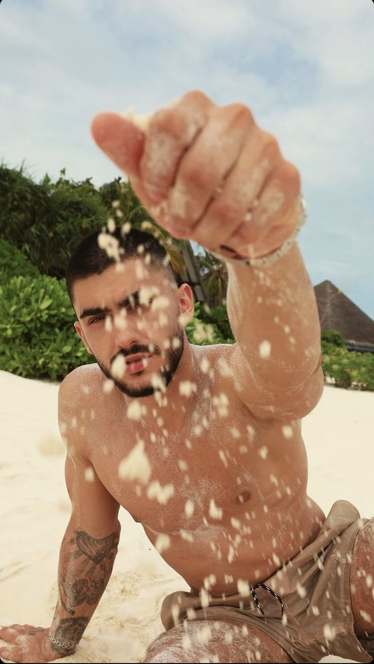 a shirtless man laying on the beach throwing sand in the air with his hand