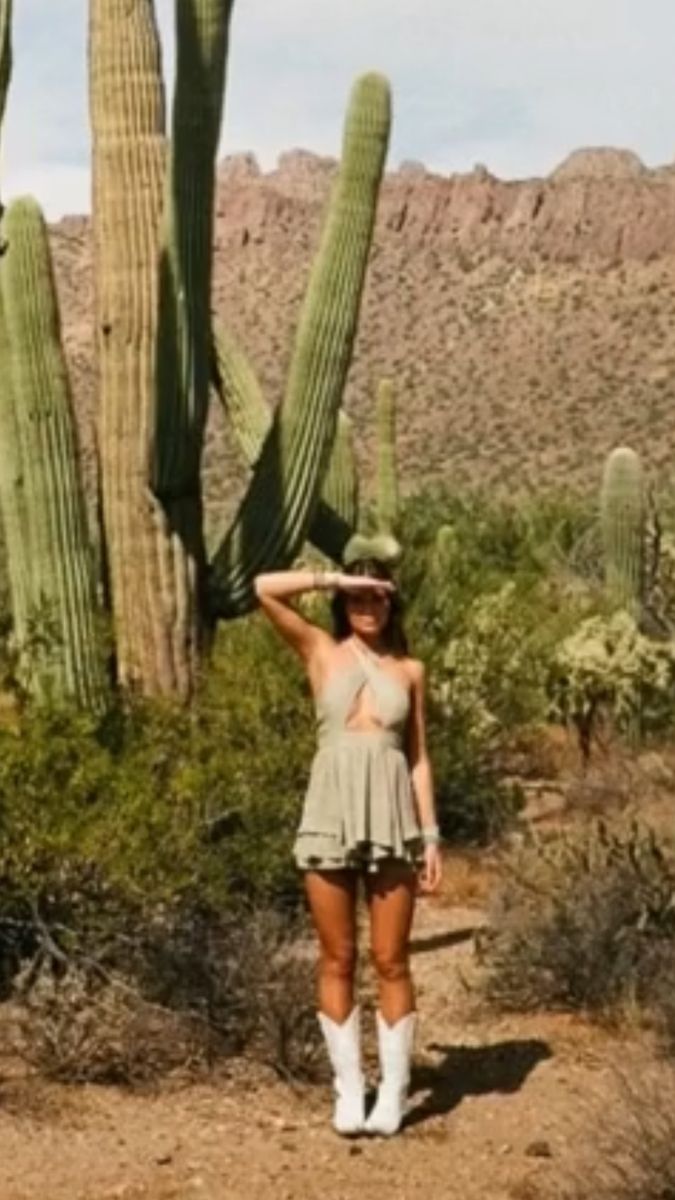 a woman standing in front of a cactus with her hands on her head and wearing white cowboy boots