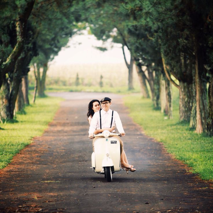 a man and woman riding on the back of a scooter down a dirt road