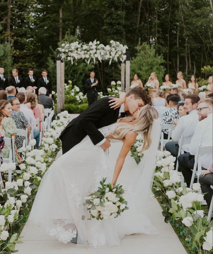 a bride and groom kissing in front of an outdoor ceremony with white flowers on the aisle