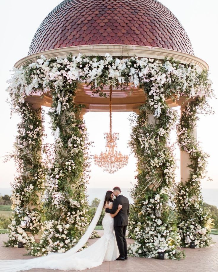 a bride and groom kissing in front of a gazebo with white flowers on it