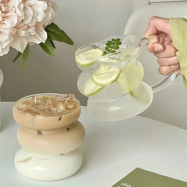 a person holding a glass filled with liquid next to a vase full of flowers and cucumbers