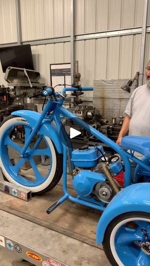 a man standing next to a blue motorcycle in a garage with other vehicles behind him