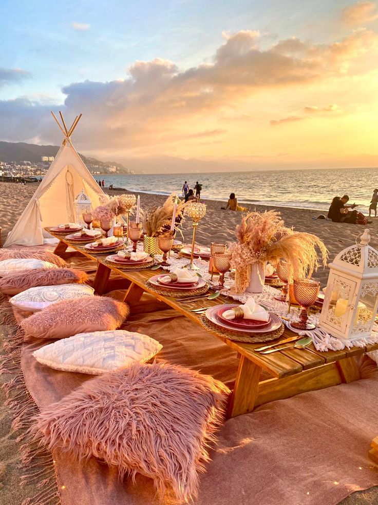 a long table set up on the beach for a party with pink pillows and blankets