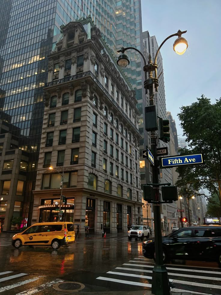 a yellow taxi cab driving down a street next to tall buildings and traffic lights on a rainy day