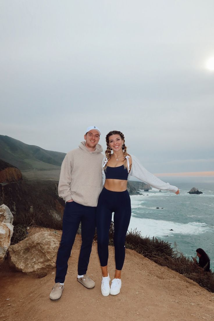 a man and woman standing on top of a hill next to the ocean