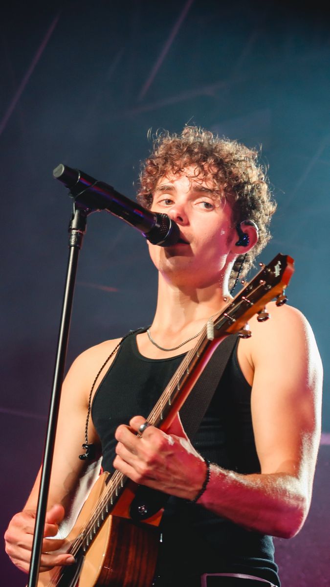 a young man holding a guitar while standing in front of a microphone on top of a stage