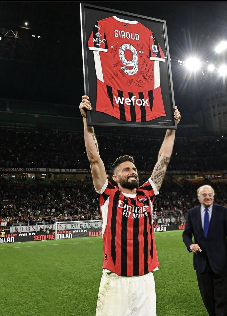 a man holding up a framed shirt on top of a soccer field with other people in the stands