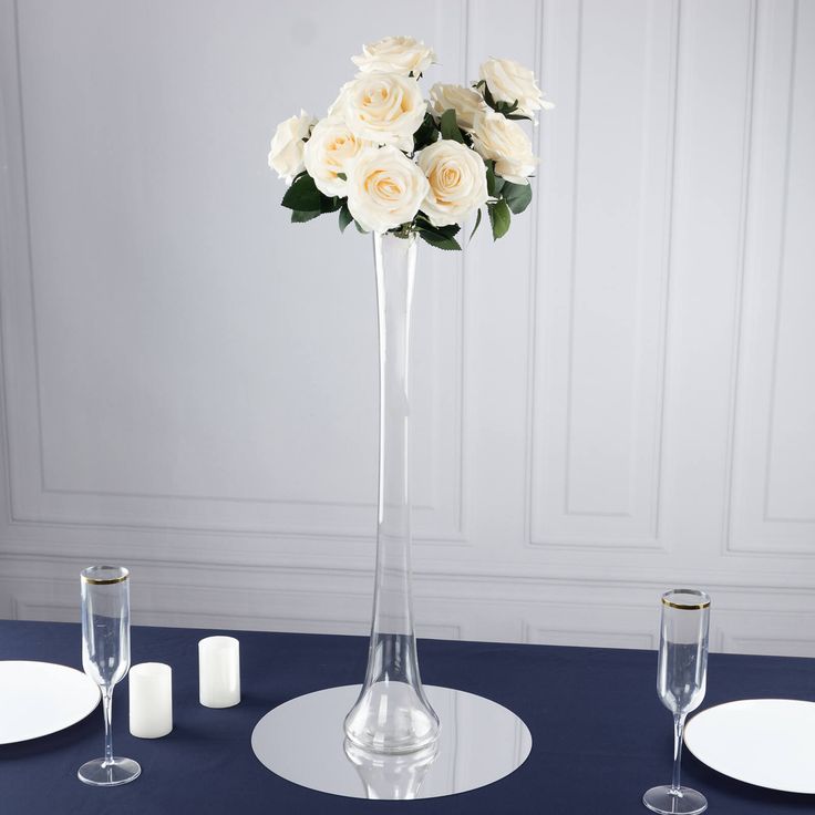 white roses in a tall vase on a blue table cloth with place settings and candles