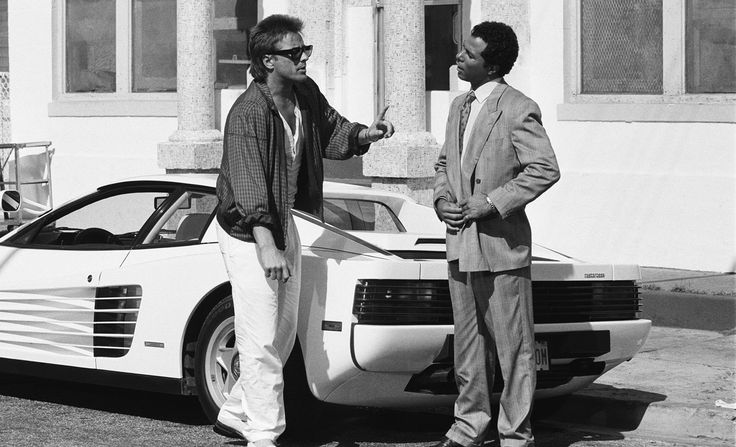 two men standing in front of a white sports car talking to each other on the street