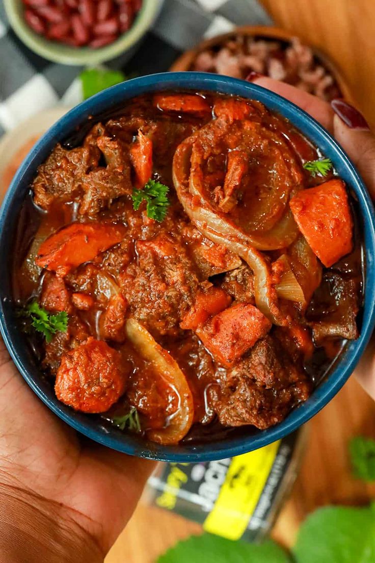 a hand holding a blue bowl filled with meat and carrots on top of a table