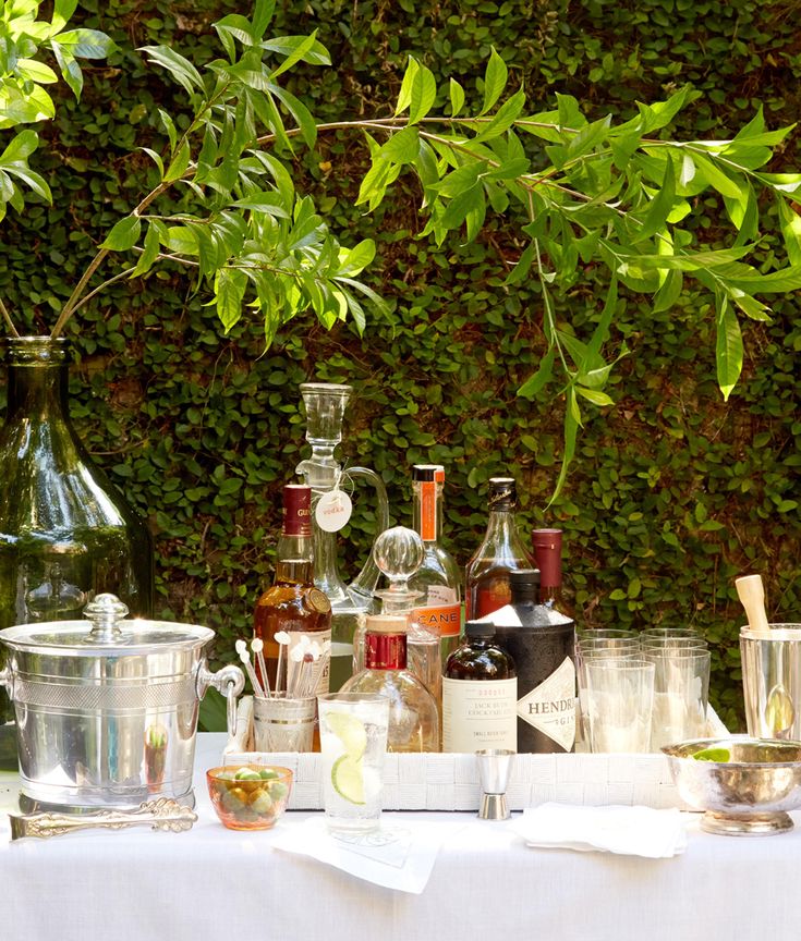 a table topped with bottles and glasses filled with liquor next to a green wall covered in ivy