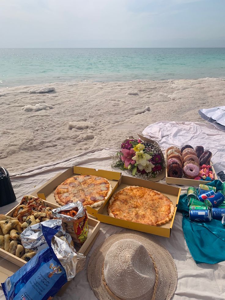 an open box of pizza on the beach with other food and snacks laid out in front of it