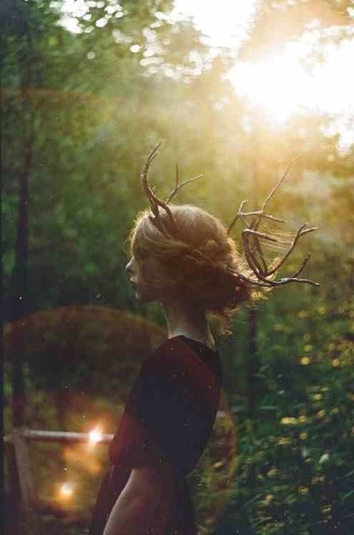 a woman with antlers in her hair is looking at the sun through some trees