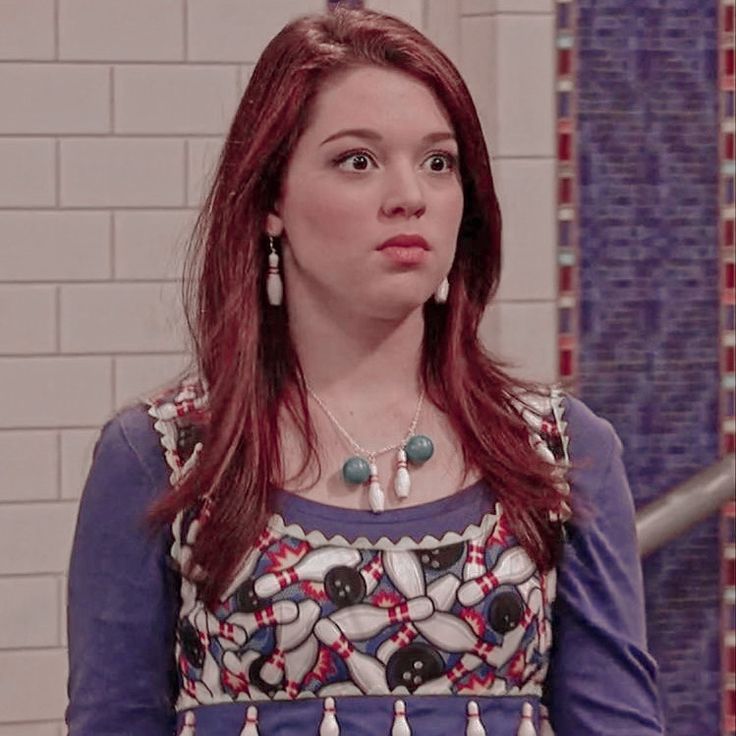 a woman with red hair wearing a blue dress and pearls on her necklace standing in front of a tiled wall