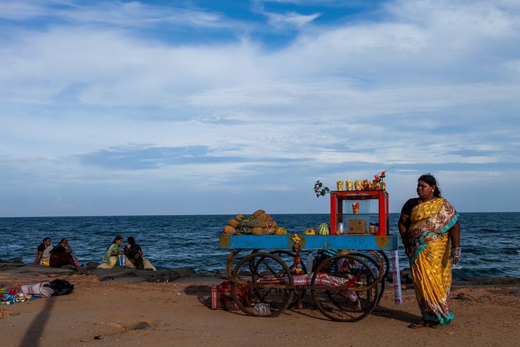 a woman standing on top of a beach next to a cart filled with fruit and vegetables