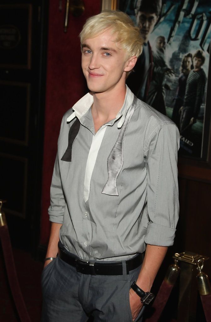 a young man with blonde hair wearing a grey shirt and black tie standing in front of a movie poster