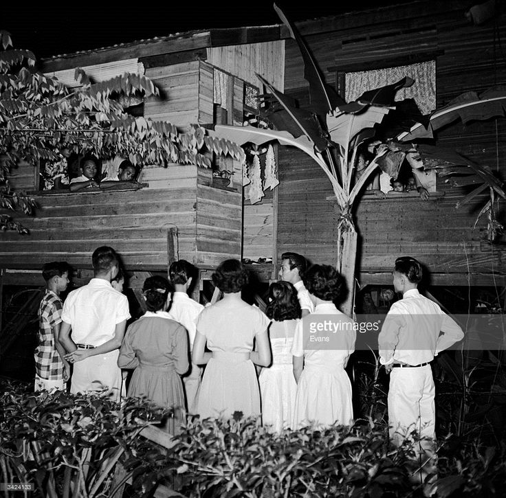 a group of people standing in front of a wooden house at night, some wearing dresses