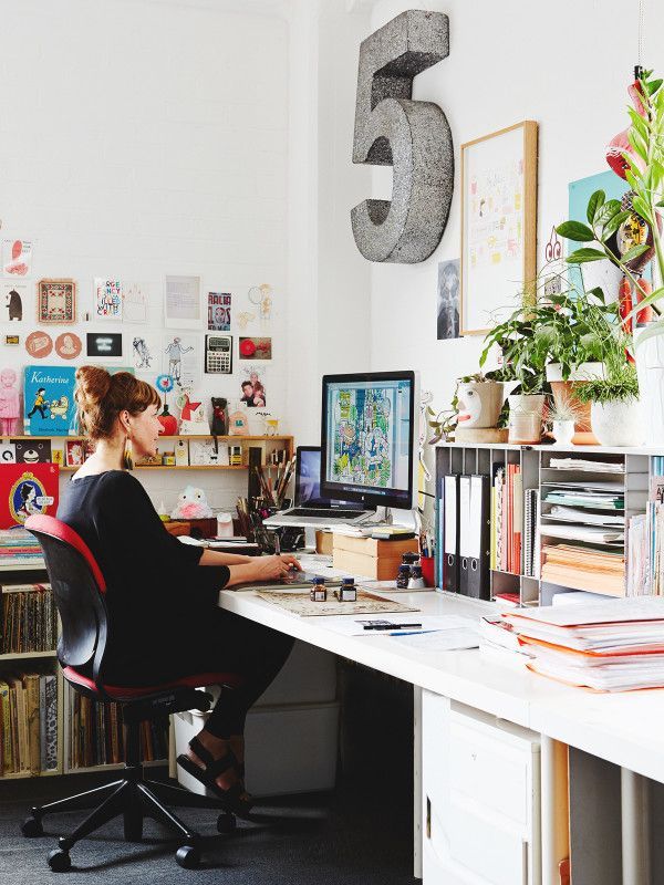 a woman sitting at a desk in front of a computer and bookshelf filled with plants