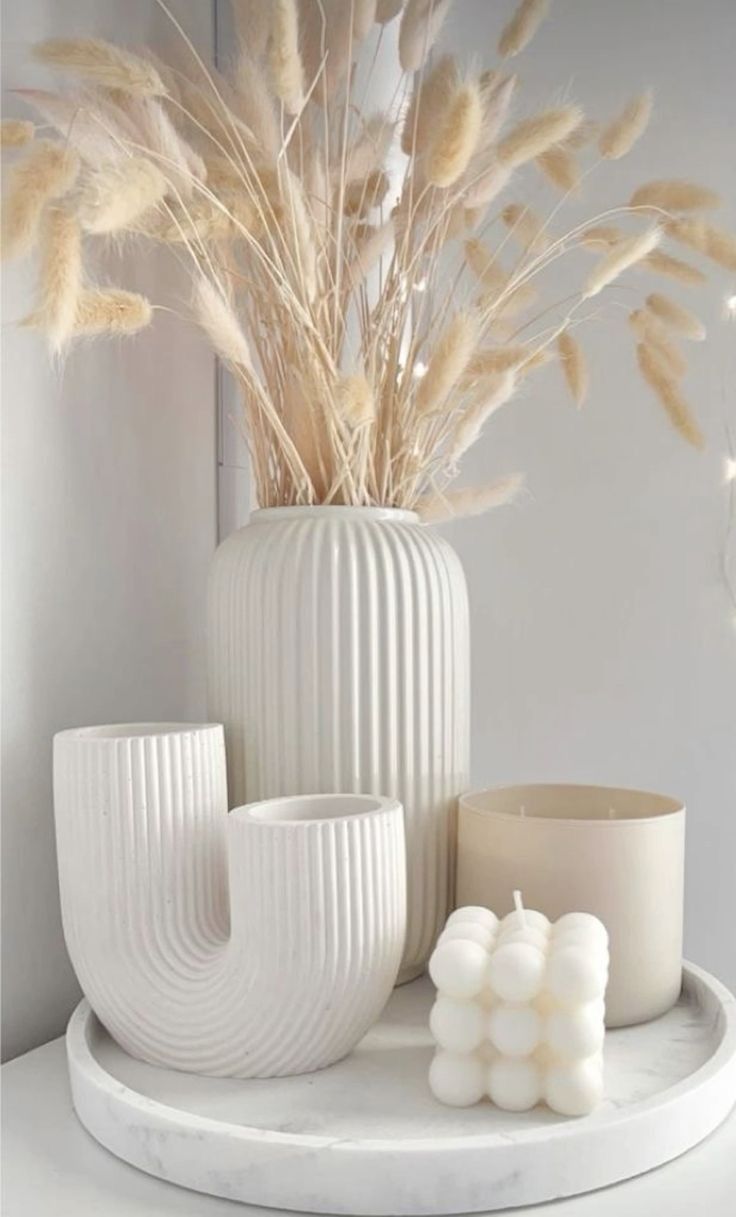 some white vases and candles are sitting on a table with dried grass in the background