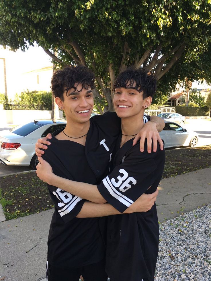 two young men standing next to each other in front of a tree and some cars