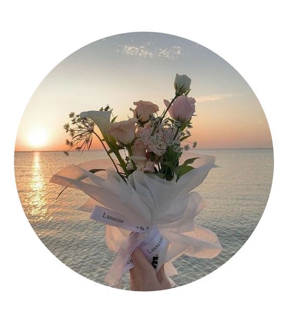 a person holding flowers in their hand on the beach at sunset or sunrise with water and sky behind them