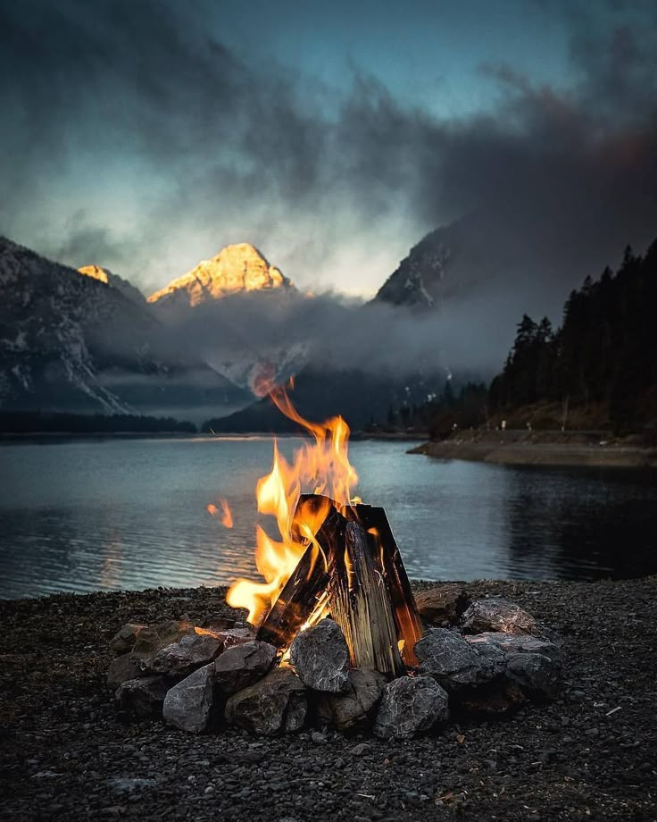 a campfire burning in the middle of a lake with mountains in the back ground