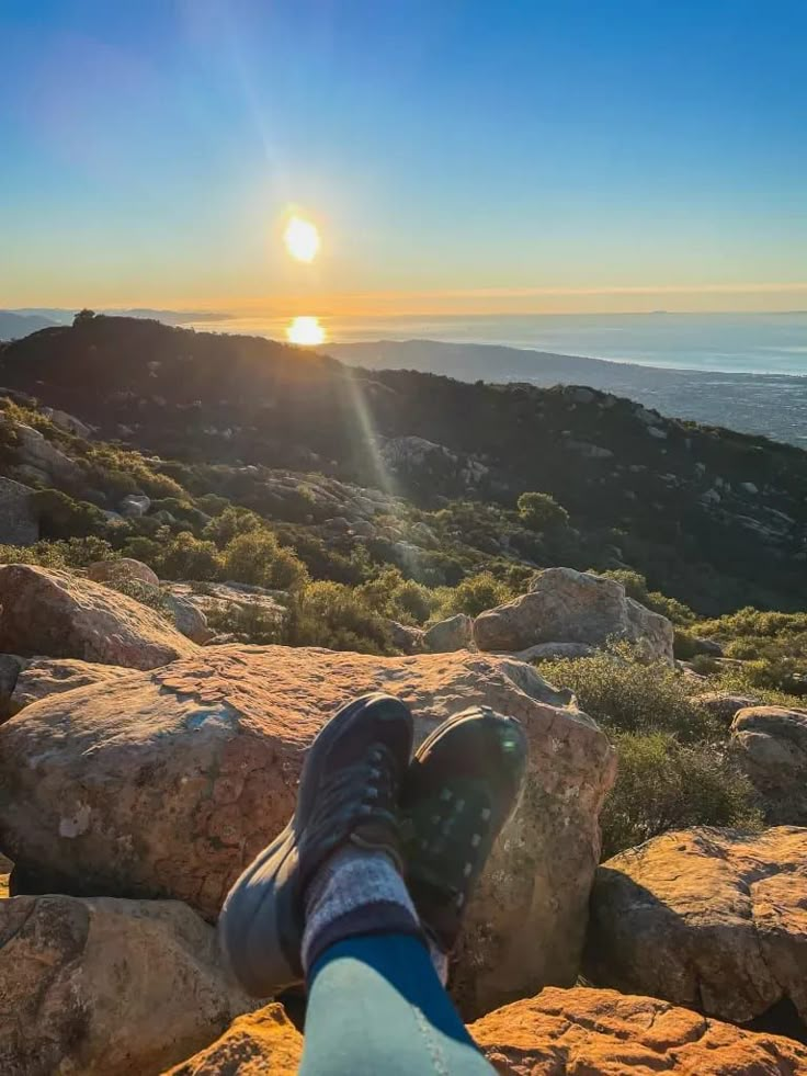 someone's feet resting on rocks overlooking the ocean and mountains at sunset or sunrise